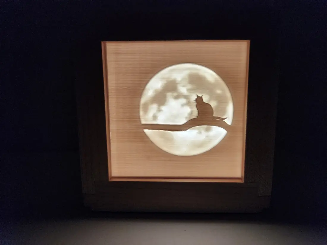 A 3D-printed light box of a cat under the moon, glowing in a wood frame